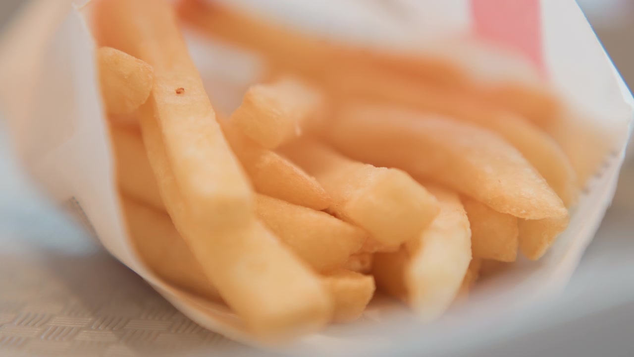 Low-angle close view showing golden french fries filling crumpled paper container, with brief finger contact lifting, shifting, and settling pieces in warm casual food scene, with fry, potato, snack