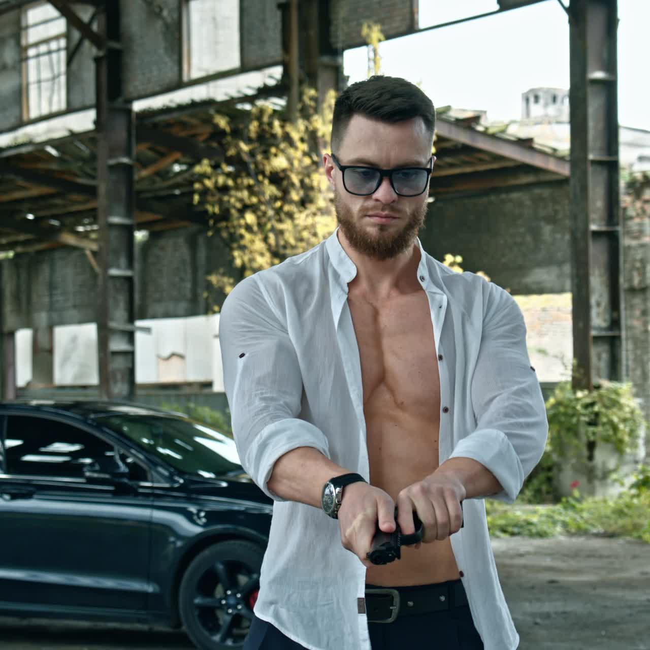 Handsome man with a pistol in ruined place. Young bearded man in glasses holds the handgun and looks at camera on luxury car background.