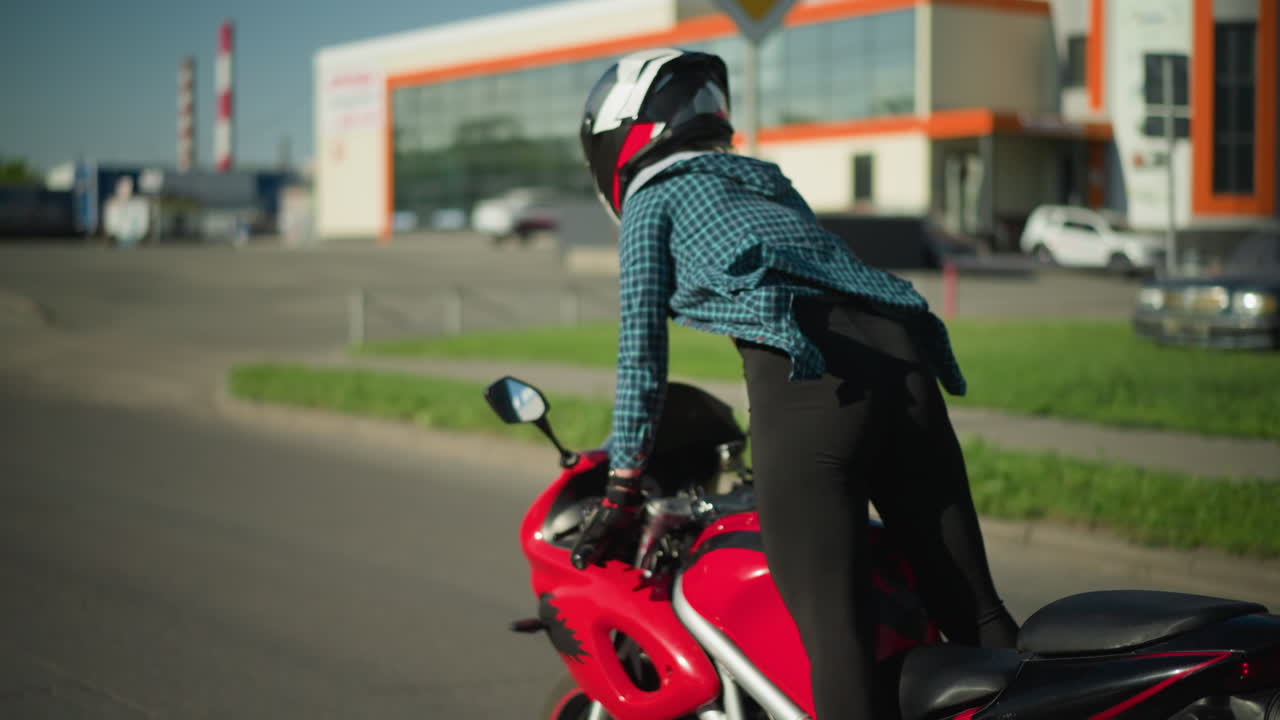 A female motorcyclist, wearing a checkered shirt and canvas shoes, rides a power bike standing, a blue truck is visible ahead of her with parked cars in the background on an open road