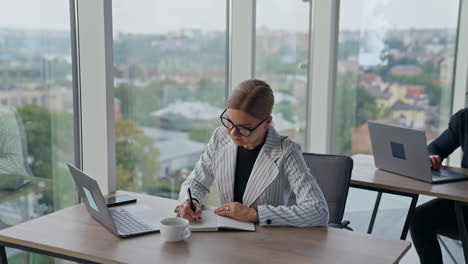 young lady writing something in her paper book. woman in glasses finishes work and takes a cup of coffee. man at backdrop works at laptop.