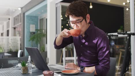 asian man with laptop drinking coffee while sitting at a cafe