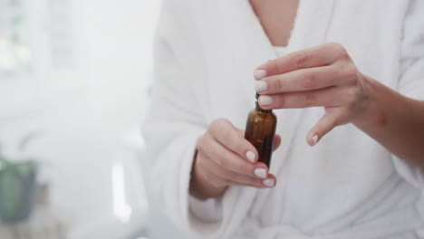 sección media de una mujer biracial aplicando aceite facial en el baño, cámara lenta