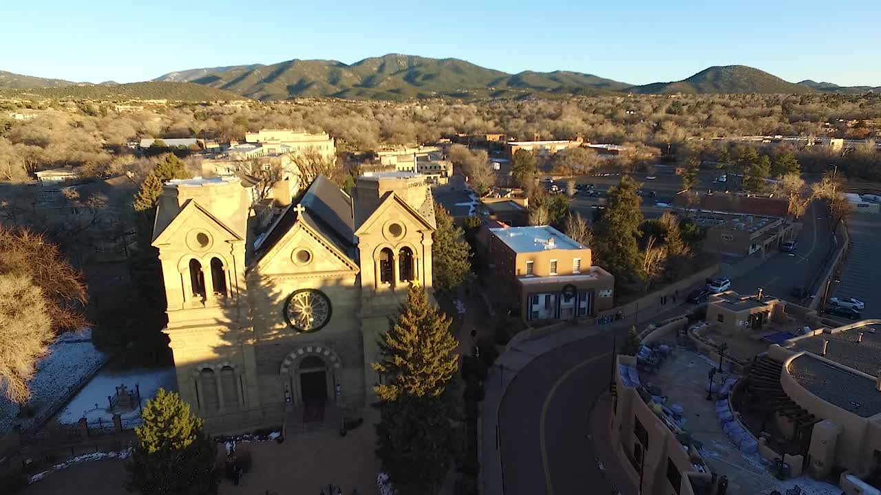 Aerial flyover of Cathedral Basilica in Santa Fe, New Mexico during golden hour in winter, with snow patches, curvy road, and mountain backdrop in warm light