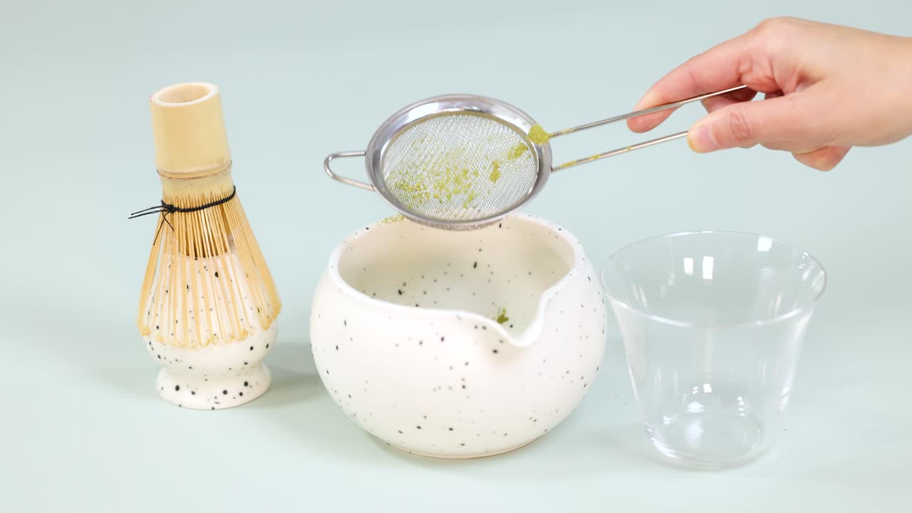 Hand sifts matcha powder through strainer into bowl, with bamboo whisk and glass cup nearby