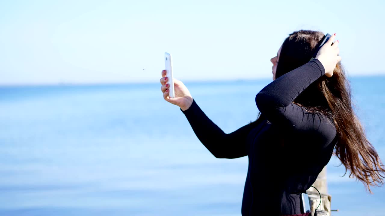 여름, 강 근처, 해변. 해가 뜨면, 아름다운 여성, 긴 어두운 머리카락, 휴대 전화, 장치에서 헤드폰에서 음악을 듣고, 전화로 비디오를 촬영, 셀피를 만니다