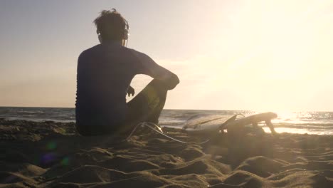 young man surfer in wetsuit sitting with surf board lying down on the sand at the beach listening to music dancing looking at the ocean at sunset