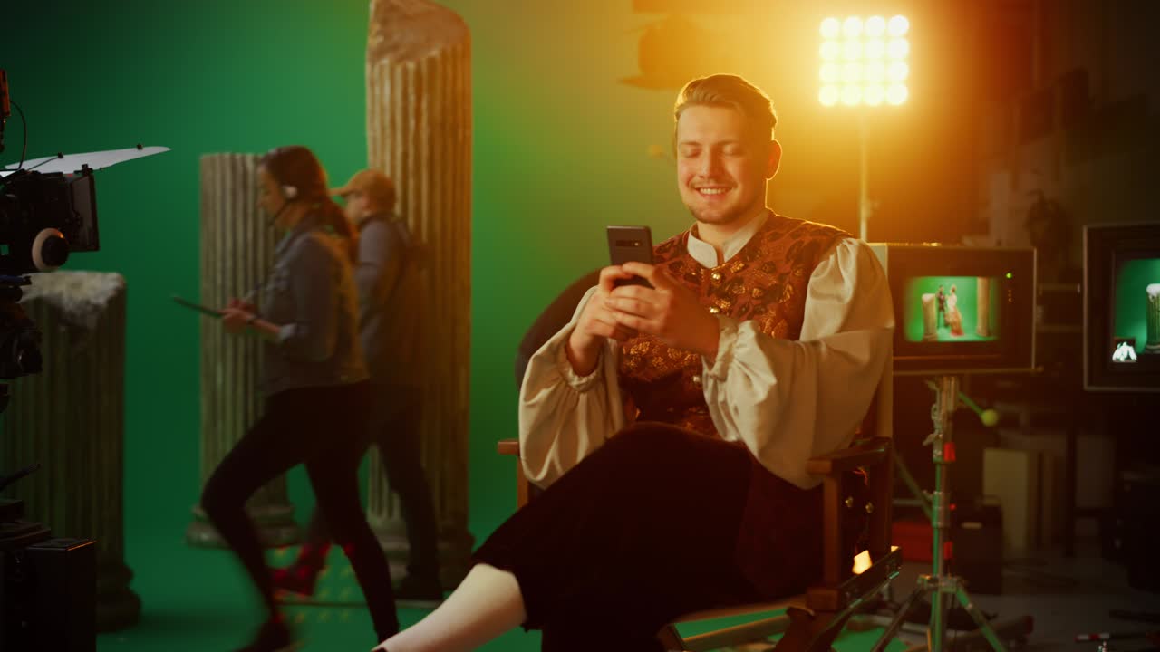 en el set de una película de drama de época: un actor guapo con un disfraz histórico sentado en una silla en un descanso usando un teléfono inteligente. en el estudio un equipo profesional filmando una película de alto presupuesto
