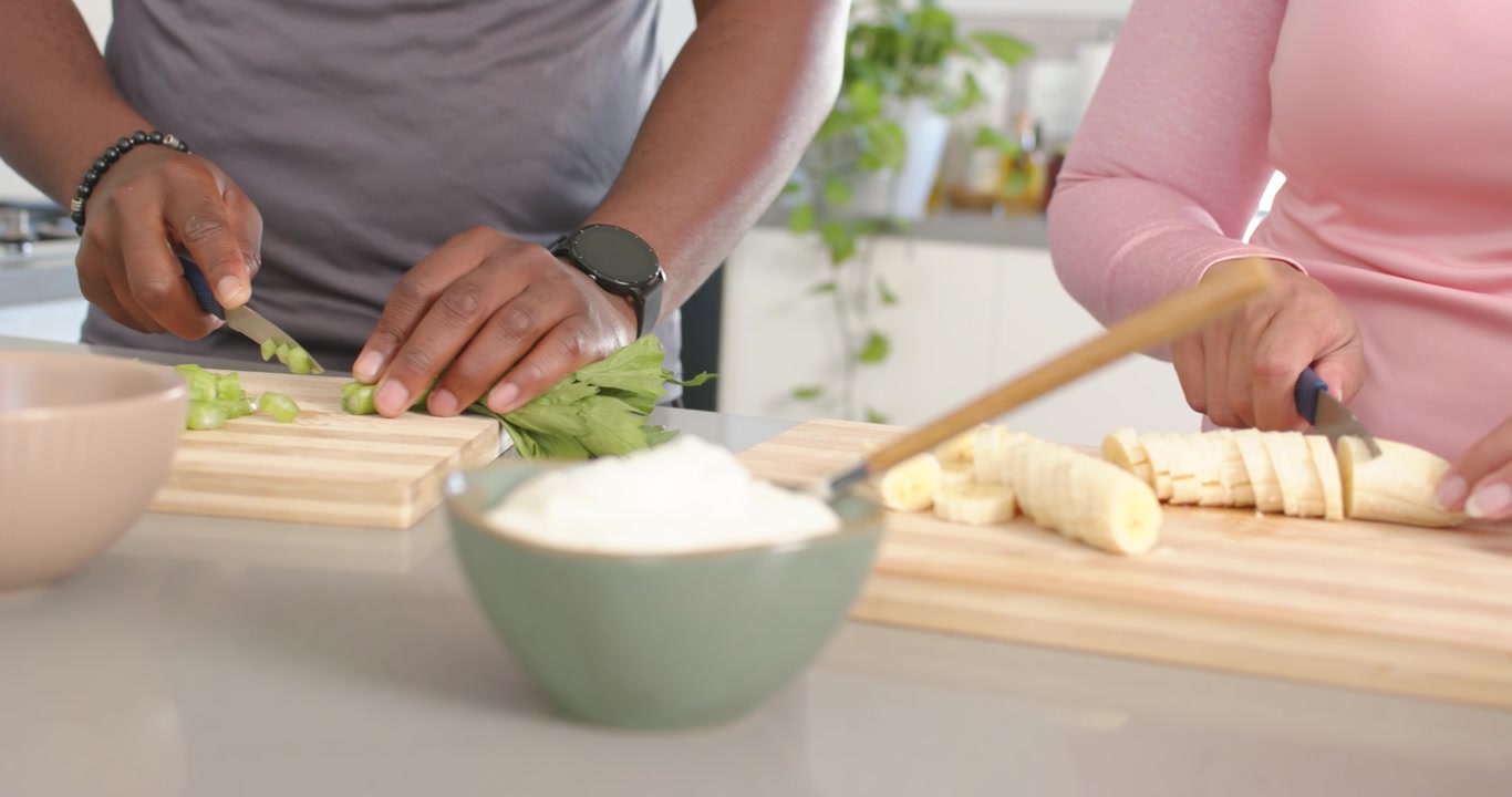 Coppia afroamericana in cucina che prepara un pasto: l'uomo inizia a tagliare il sedano, la donna taglia la banana.