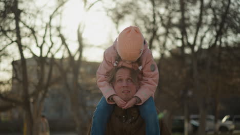 a little girl in a pink cap and jacket sits joyfully on her dad's neck as they walk through a sunlit park. the girl bends her head down while the dad looks up at her