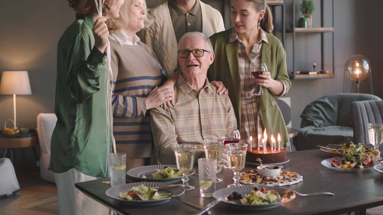 Senior Man Blowing Out Candles On Birthday Cake During A Celebration With His Family At Home
