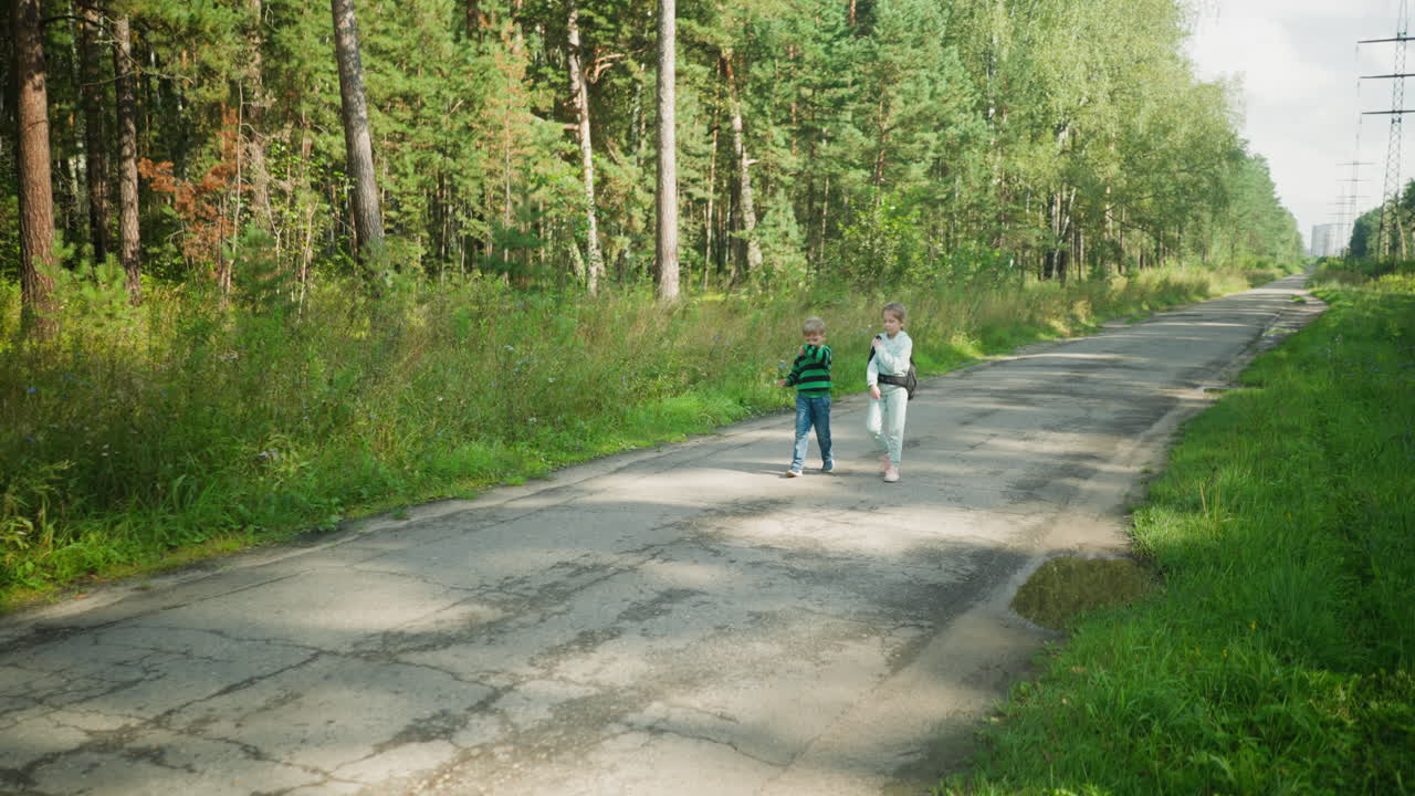 Two children walking home side by side on cracked tarred road surrounded by tall forest trees and green vegetation, with water puddle nearby and sunlight casting soft shadows on countryside path