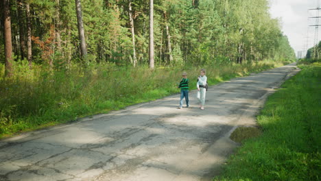 two children walking home side by side on cracked tarred road surrounded by tall forest trees and green vegetation, with water puddle nearby and sunlight casting soft shadows on countryside path