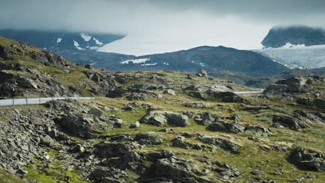 a winding mountain road cuts through a rugged, rocky landscape, with snow-capped mountains and a glacier visible in the distance