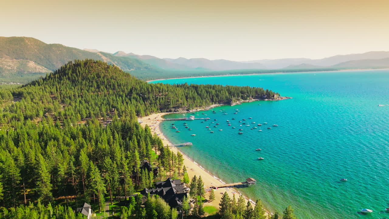 Lovely view of wonderful blue Lake Tahoe with boats at the shore. Sunny sandy beach and pine tree forest at waterfront. Hazy mountains at backdrop.