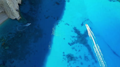 Tourists-ride-boats-to-Navagio-Beach-cove-along-the-coast-of-Zakynthos-Island,-Greece---aerial-reveal