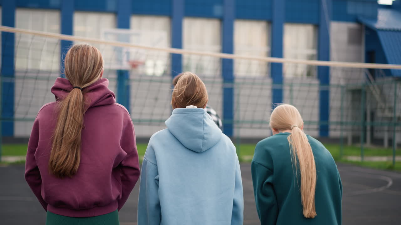 다양한 색의 후디를 입은 젊은 운동선수들이 야외 코트, 네트, 파란색 건물, 그리고 배경에 보이는 초록색에서 움직이고 있는 체크색 셔츠를 입은 배구 강사를 관찰하는 뒷면