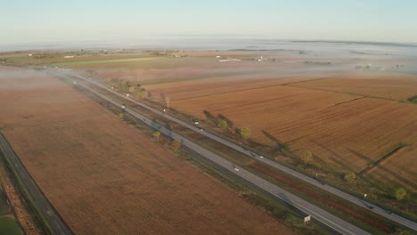 Morning-aerial-view-of-rural-highway-surrounded-by-orange-agricultural-fields-with-thin-layer-of-early-fog-hanging-over-the-landscape