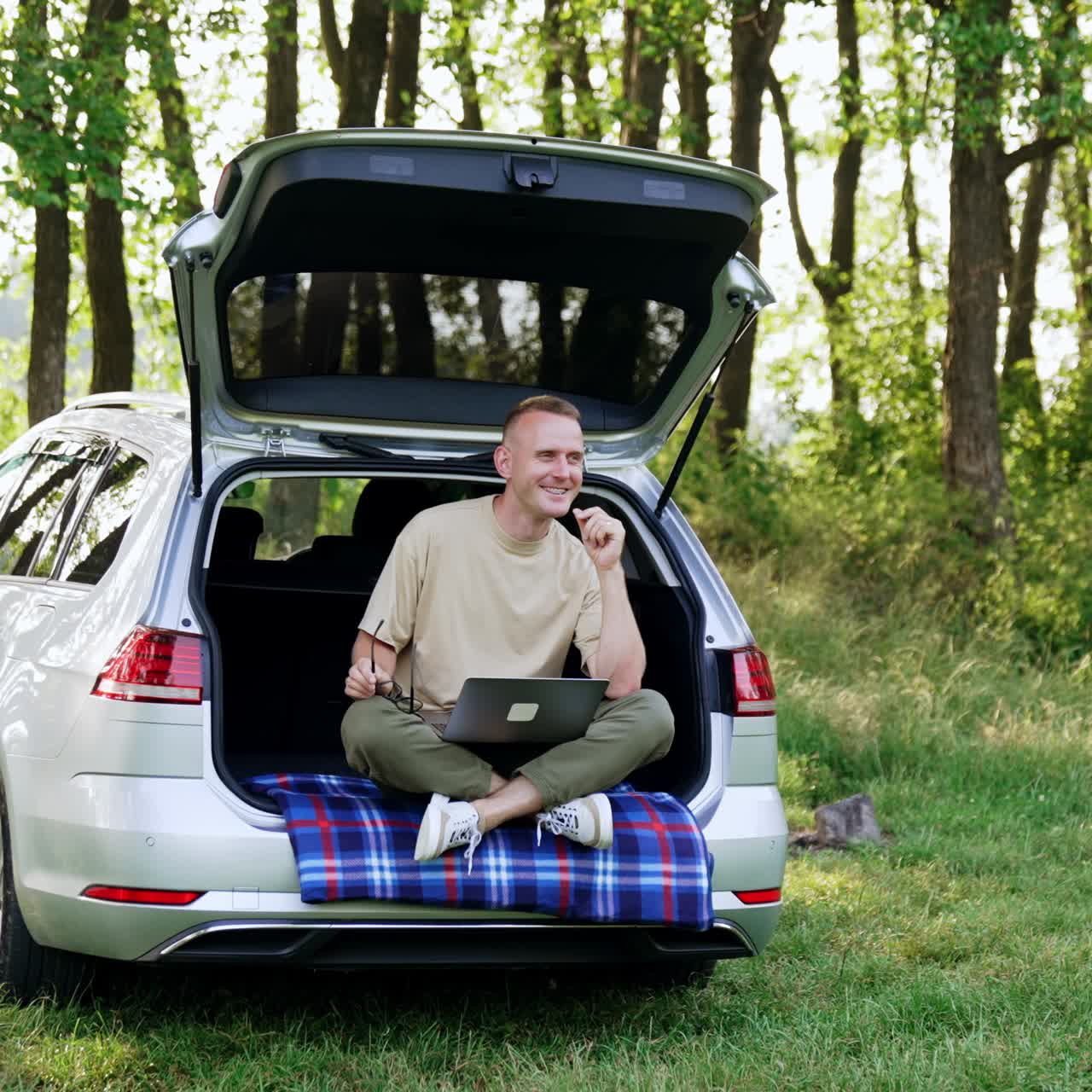 Thoughtful Caucasian man works in nature on his laptop. Happy man sits in car trunk looking pensively around, thinking about ideas