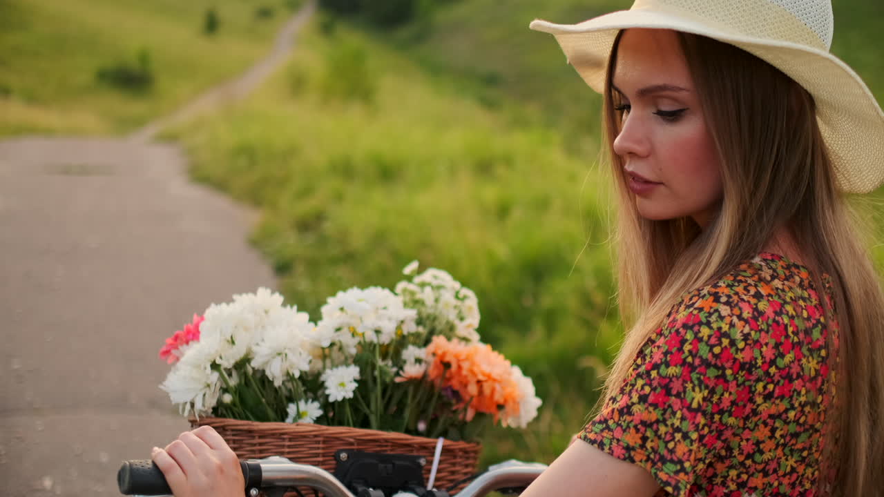 una chica rubia feliz con vestido y sombrero se da la vuelta y sonriendo alegremente mira a la cámara y coquetea paseando por el campo en verano con bicicleta y flores.