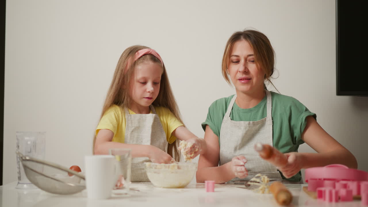 Woman in green shirt adjusts cup and rolling pin while teaching young girl how to knead dough during baking activity, both wearing aprons and focused on preparation with tools on white table