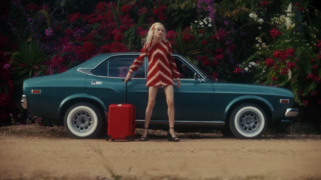 Donna che cammina con una valigia rossa accanto a un'auto verde vintage