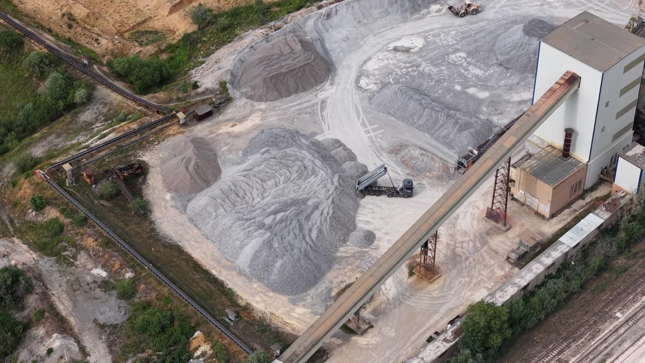 An aerial view captures a quarry operation with a truck dumping gravel near a large industrial building and conveyor system.