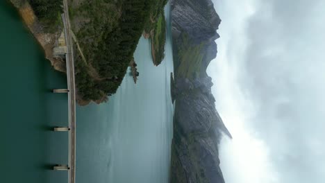 Aerial-Drone-Shot-of-Riaño-Lake-in-Picos-de-Europa,-Spain,-with-Cloudy-Skies,-Calm-Blue-Waters,-Mountain-Cliffs,-and-Lush-Green-Trees