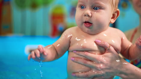 chubby caucasian baby is in the swimming pool. female coach moves the kid in water and he splashes it happily.