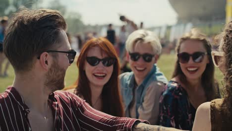 group of friends sitting on grass together at music festival and drinking beer while chatting.