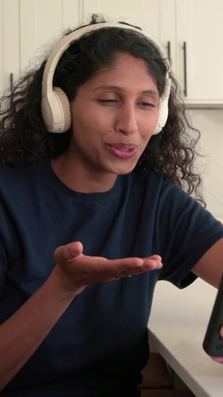 Vertical video: Waving woman in navy shirt, white headset on call at kitchen counter holding phone