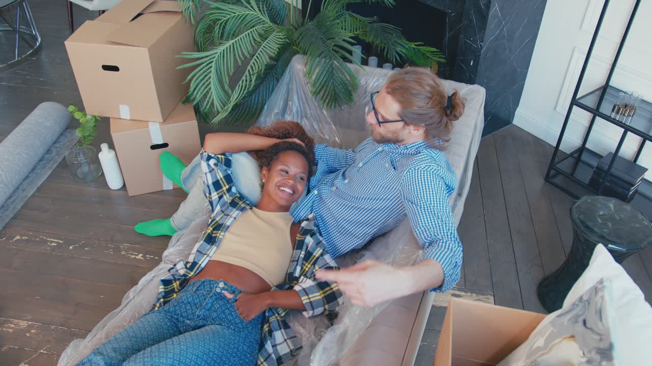 African american woman lies on lap of caucasian man sits on sofa after moving