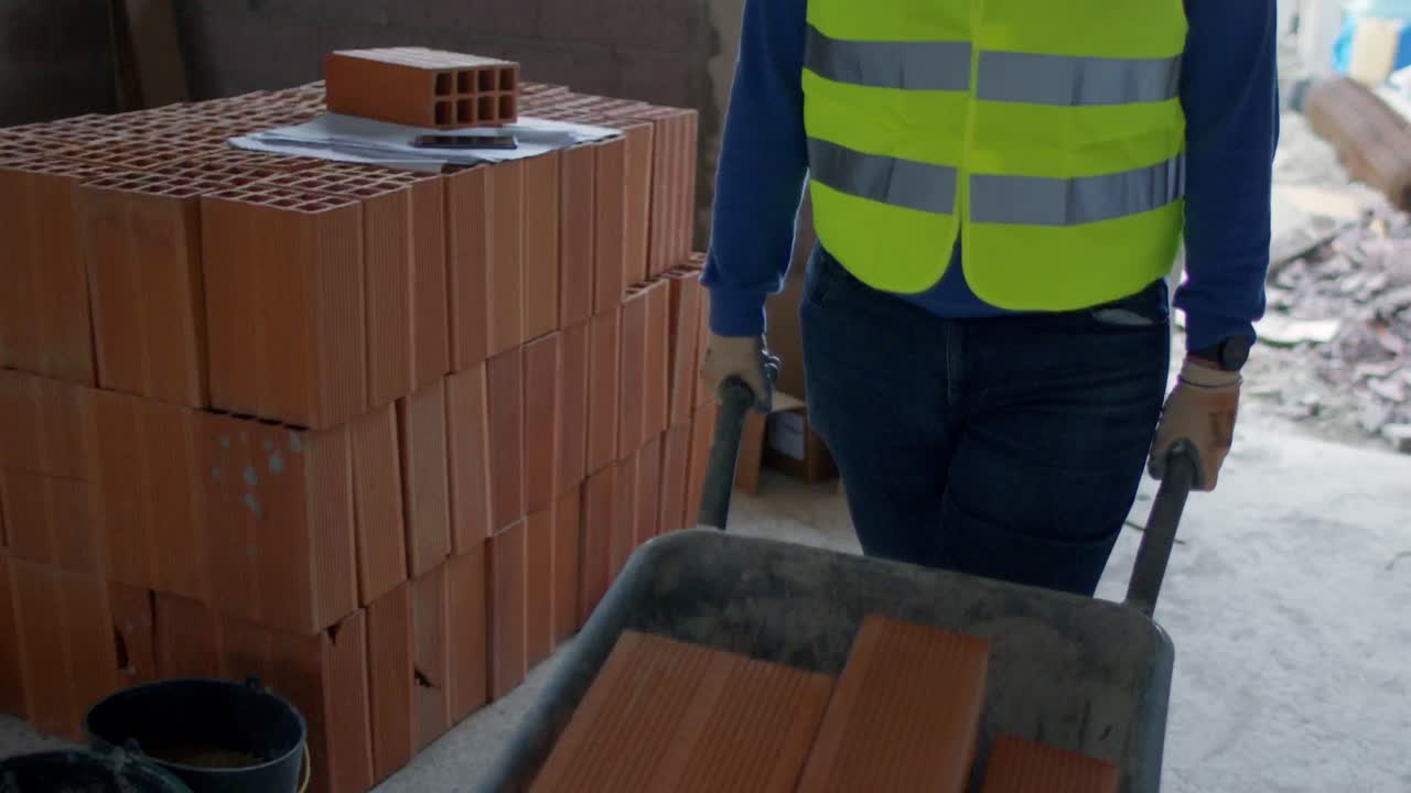 4K slow-motion footage of a construction worker in a safety vest pushing a wheelbarrow filled with bricks at a building site, showcasing labor and effort.
