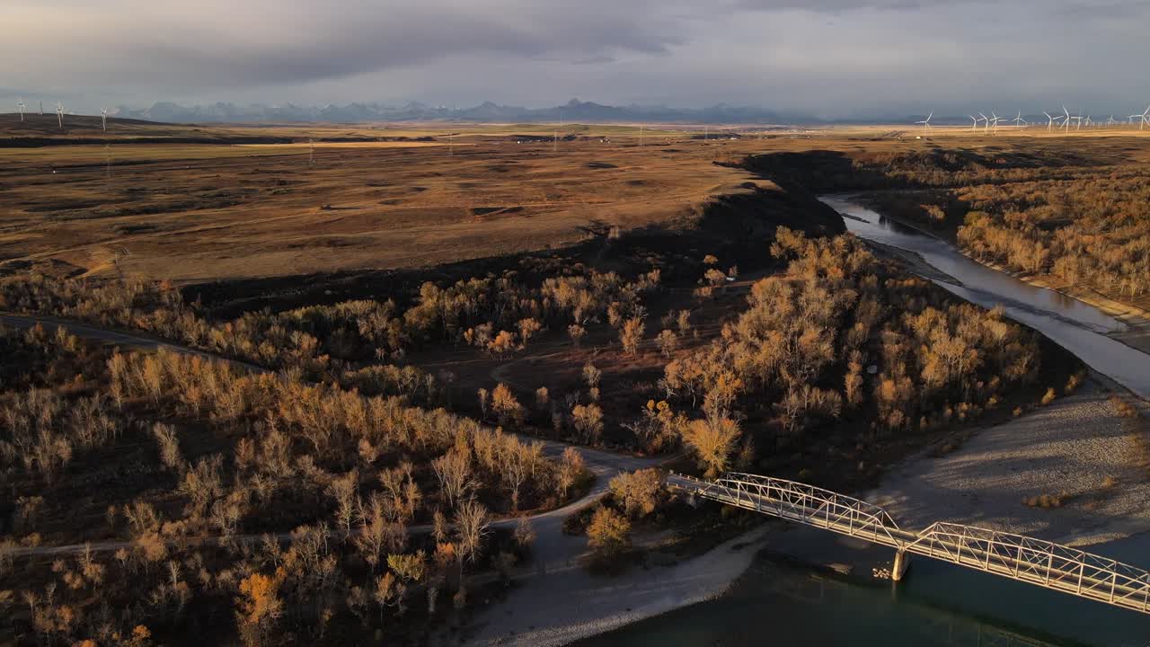 4k aerial footage approaching truss bridge near Brocket, Alberta. Southern Kootenays in background underneath heavy clouds. Sunrise gloom causing warm tones. Cantilever bridge crossing old man river.