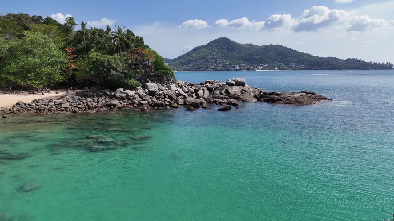 Drone footage captures the serene coastline of Phuket, Thailand, showcasing turquoise waters, lush greenery, and rocky shores under clear skies