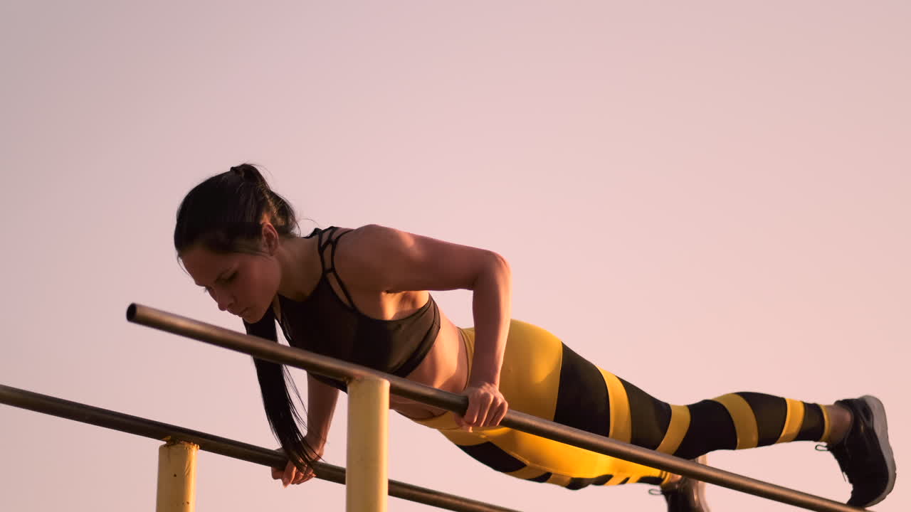 cámara lenta: una hermosa atleta haciendo flexiones en barras con una camiseta negra en gran forma y pantalones amarillos con cabello oscuro.