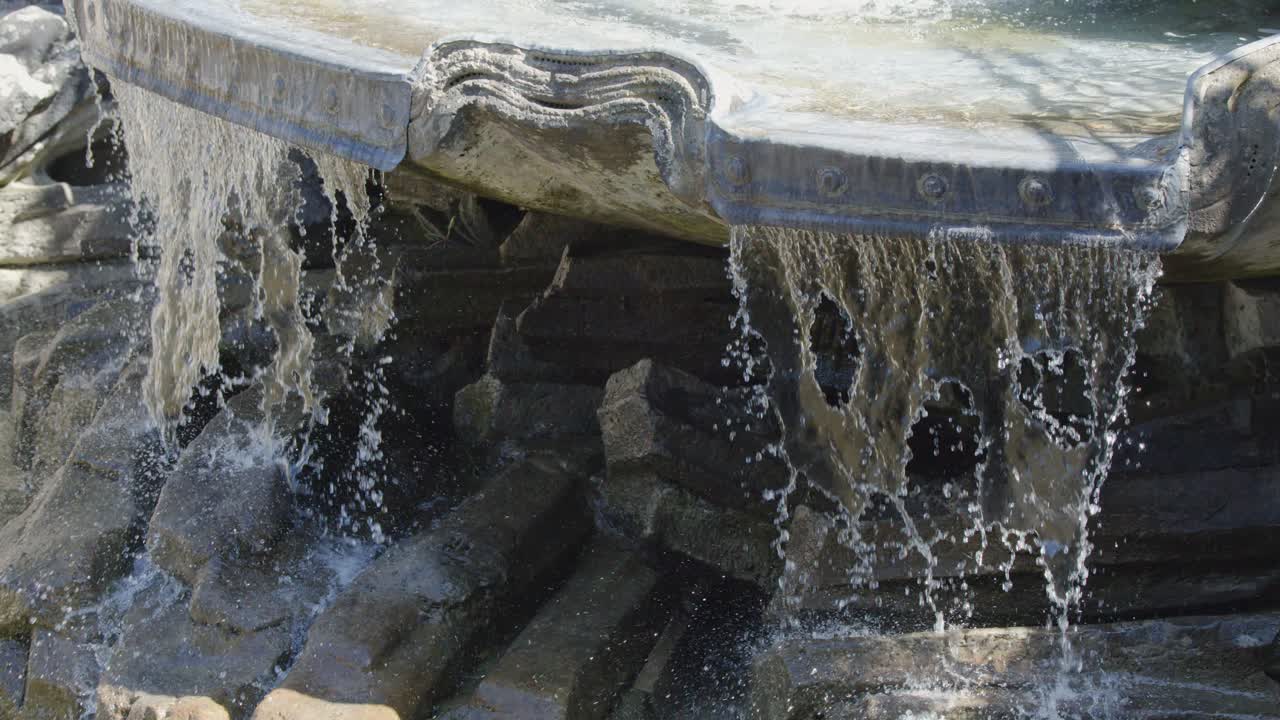 Clear water cascades over sculpted stone fountain, splashing onto rocks in bright natural daylight
