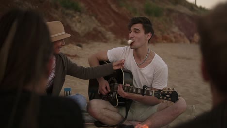 Pareja-Joven-Romántica-Sentada-Junto-Al-Fuego-En-La-Playa-Junto-Con-Amigos