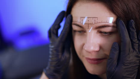 cosmetologist in black gloves attaches liner on the female client's forehead. procedure of eyebrows permanent make up. close up.
