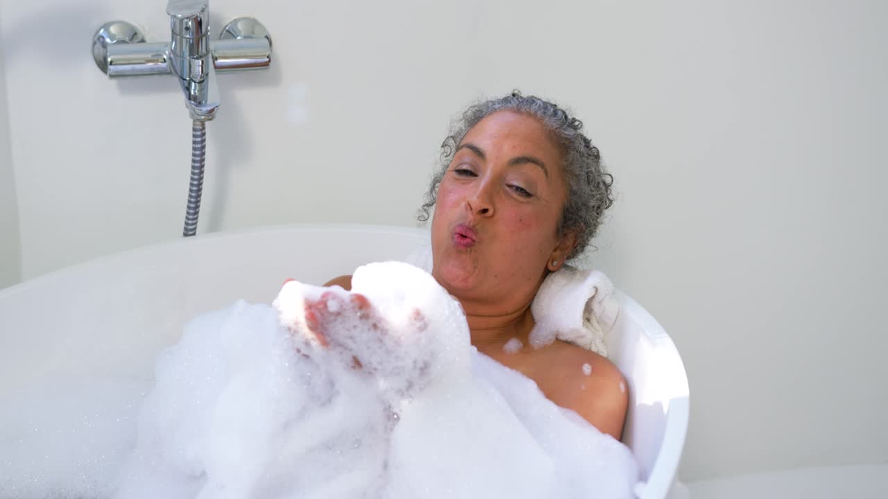 Senior African American woman reclining in tub cupping foam then blowing bubbles for calm