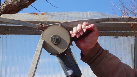 hands with an angle grinder cut an old metal greenhouse against a blue sky.