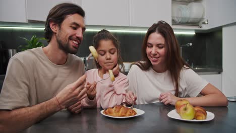 Un-Hombre-Moreno-Feliz-Con-Barba-Incipiente-Con-Una-Camiseta-Beige-Junto-Con-Su-Esposa-Morena-Con-Una-Camiseta-Blanca-Y-Una-Pequeña-Hija-Con-Ropa-Rosa-Están-Desayunando-En-Un-Apartamento-Moderno-En-Una-Mesa-De-Comedor-Negra-Por-La-Mañana.