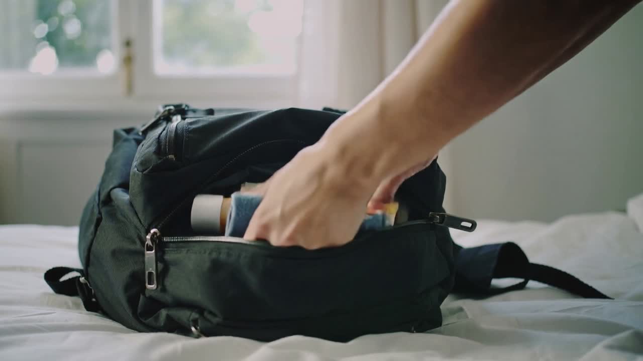 Tangan masuk dari kanan mengemas pakaian dan perlengkapan mandi ke dalam saku ransel hitam di tempat tidur untuk perjalanan.