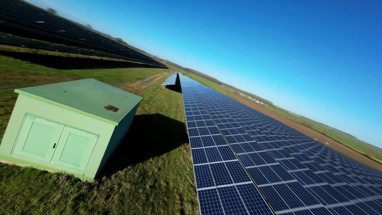 Dynamisk lavpas FPV-flyvning, der suser forbi rene energianlæg placeret på åbne grønne marker i det tidlige morgenlys.