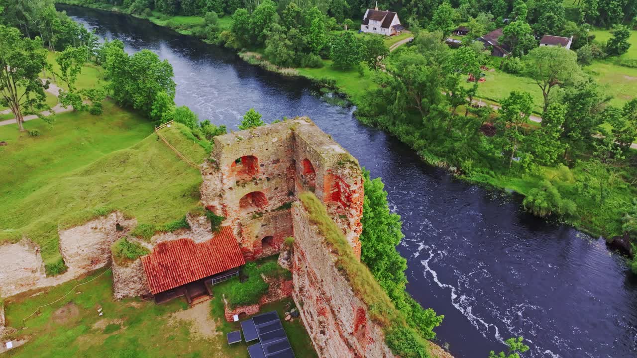 Drone of ruined tower of Bauska Castle beside Memele River as bird glides by