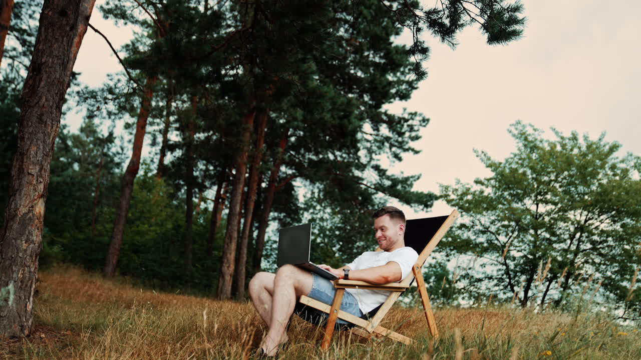 Happy Caucasian man working remotely. Businessman in t-shirt and jeans sits in a folding chair holding laptop. Nature at backdrop. Low angle view.