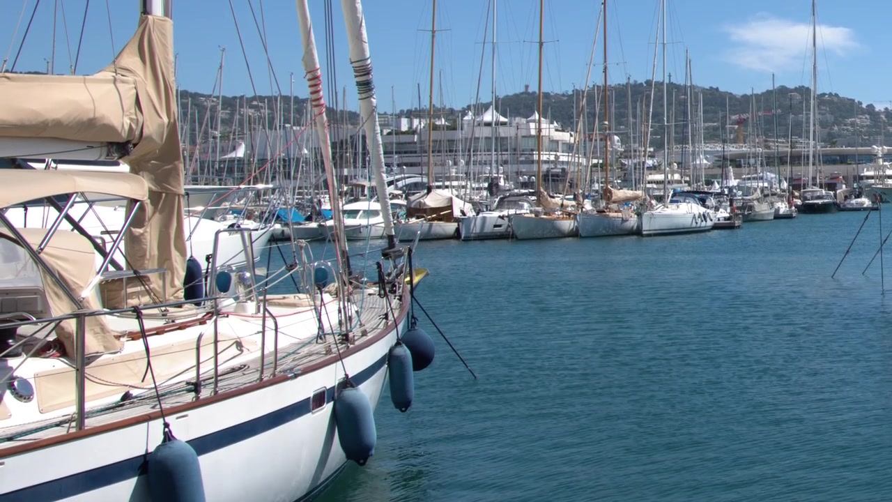 Inquadratura ampia e statica di una barca a vela bianca ormeggiata in un affollato porto della Costa Azzurra.