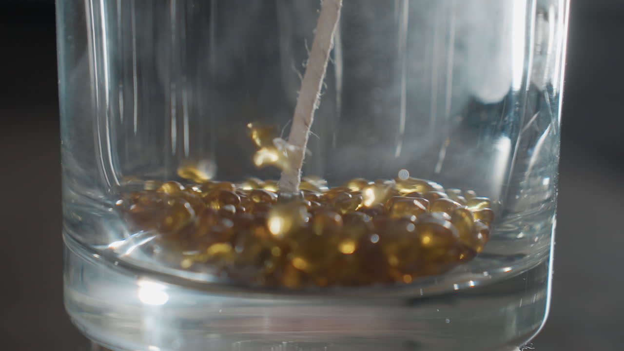 Close up of transparent glass cup with vertical wick as golden brown decorative beads fall rapidly inside, scattering across shiny base, creating vibrant shimmer and motion effect