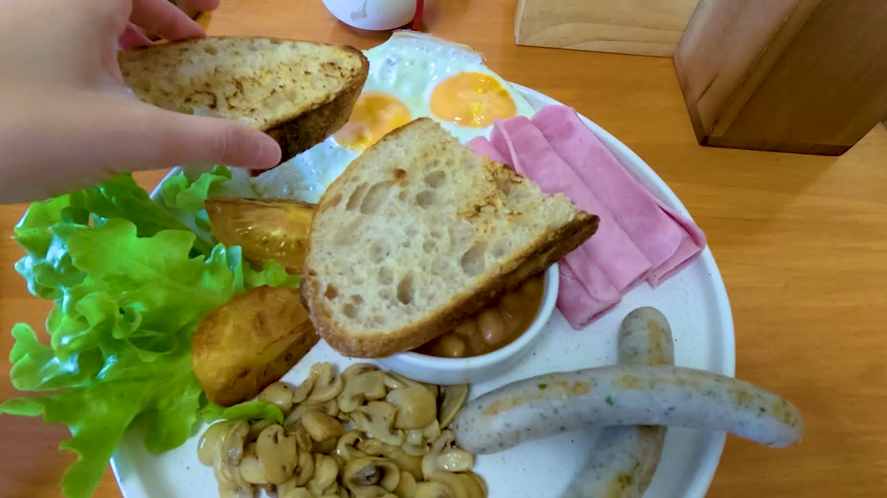 A hand arranges bread on a breakfast plate with baked beans, sausages, and greens. Bright lighting enhances the vibrant colors