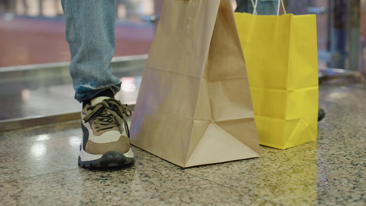 Close up of person wearing sneaker standing beside shopping bags on shiny mall floor with casual jeans reflecting light modern retail lifestyle urban consumer