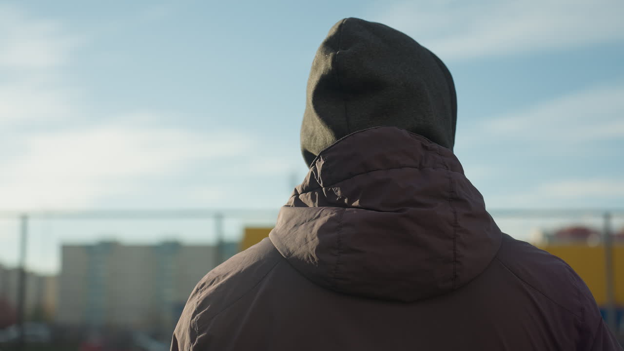 후디와 재을 입은 남자가 도시 스포츠 경기장에서 주변을 둘러보며, 주거 건물의 흐릿한 배경, 활기찬 노란색 벽, 밝은 빛으로 조명됩니다.